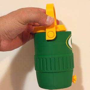 Puede incluir: Nevera aislante verde y amarilla con asa. La nevera tiene forma cil&iacute;ndrica con un dise&ntilde;o acanalado en el centro. Un asa amarilla est&aacute; unida a la parte superior y una boquilla amarilla en el lateral. Un logotipo es visible en el lateral.