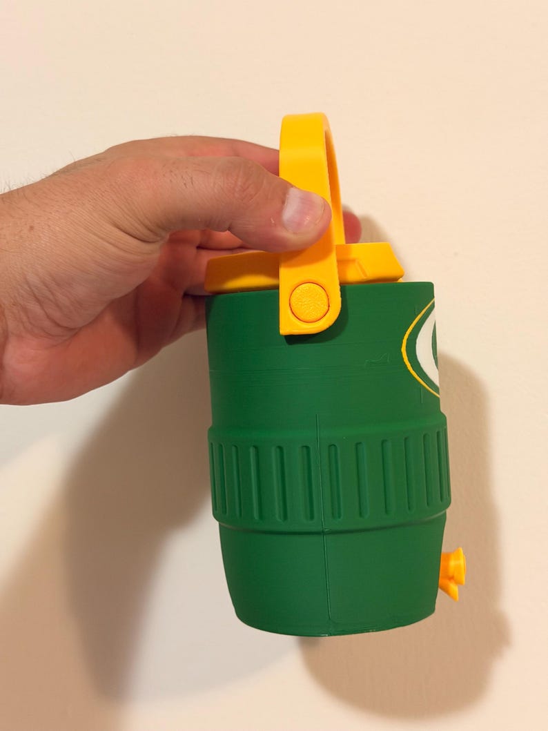 Puede incluir: Nevera aislante verde y amarilla con asa. La nevera tiene forma cil&iacute;ndrica con un dise&ntilde;o acanalado en el centro. Un asa amarilla est&aacute; unida a la parte superior y una boquilla amarilla en el lateral. Un logotipo es visible en el lateral.