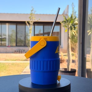 May include: A blue and yellow insulated water cooler with a stainless steel straw. The cooler has a yellow handle and a small yellow spout. The cooler is on a black surface, with a house and trees in the background.