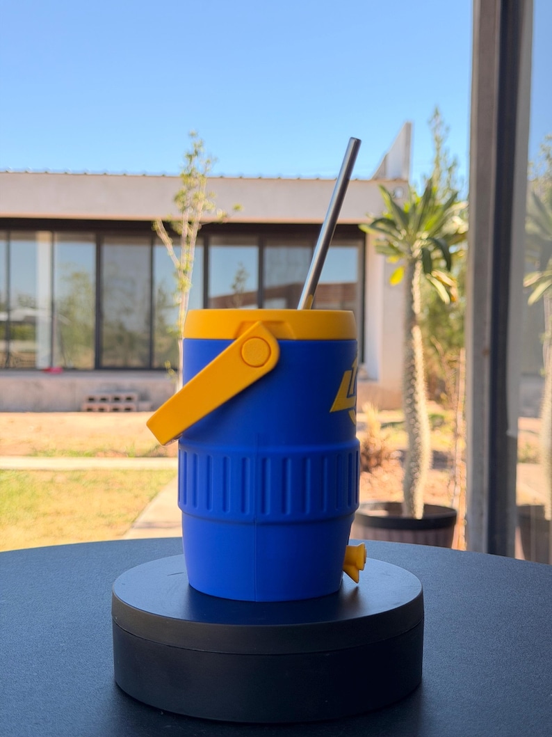 May include: A blue and yellow insulated water cooler with a stainless steel straw. The cooler has a yellow handle and a small yellow spout. The cooler is on a black surface, with a house and trees in the background.