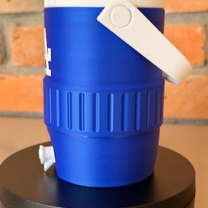 May include: A blue and white water cooler with a white handle and a spigot. The cooler has the letters "LA" in white on the side. The cooler is sitting on a black surface.