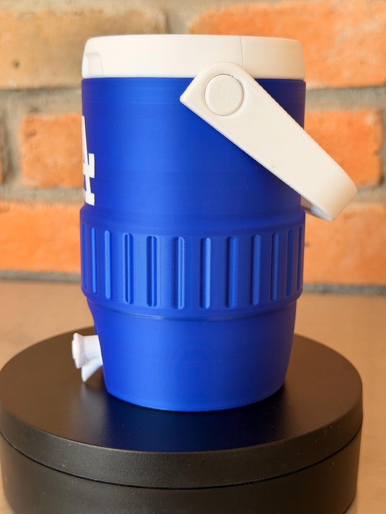 May include: A blue and white water cooler with a white handle and a spigot. The cooler has the letters "LA" in white on the side. The cooler is sitting on a black surface.