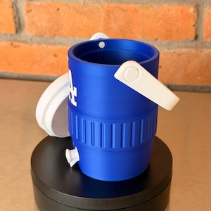 May include: A blue and white miniature bucket with the Los Angeles Dodgers logo. The bucket has a white handle and a white lid. The bucket is sitting on a black surface in front of a brick wall.