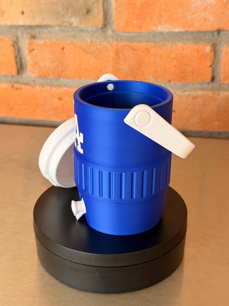 May include: A blue and white miniature bucket with the Los Angeles Dodgers logo. The bucket has a white handle and a white lid. The bucket is sitting on a black surface in front of a brick wall.