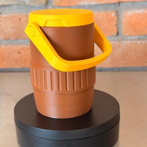 May include: A brown and yellow cooler with a handle. The cooler has a cylindrical shape with a ribbed base and a yellow lid. The handle is also yellow. The cooler is sitting on a black circular base.