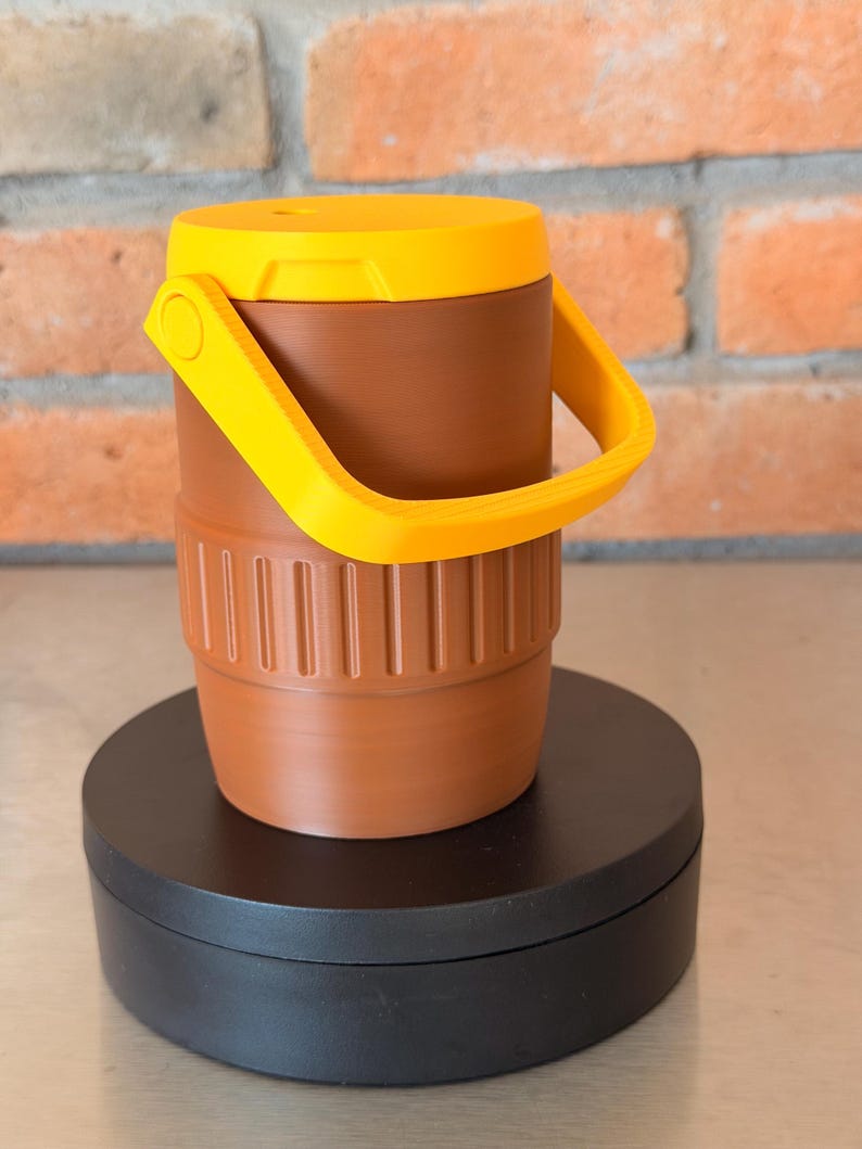 May include: A brown and yellow cooler with a handle. The cooler has a cylindrical shape with a ribbed base and a yellow lid. The handle is also yellow. The cooler is sitting on a black circular base.