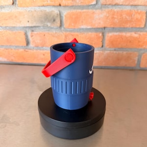 May include: A navy blue bucket-shaped container with a red handle and accents. The container has a white logo on the side and is sitting on a black circular base. The background is a brick wall.
