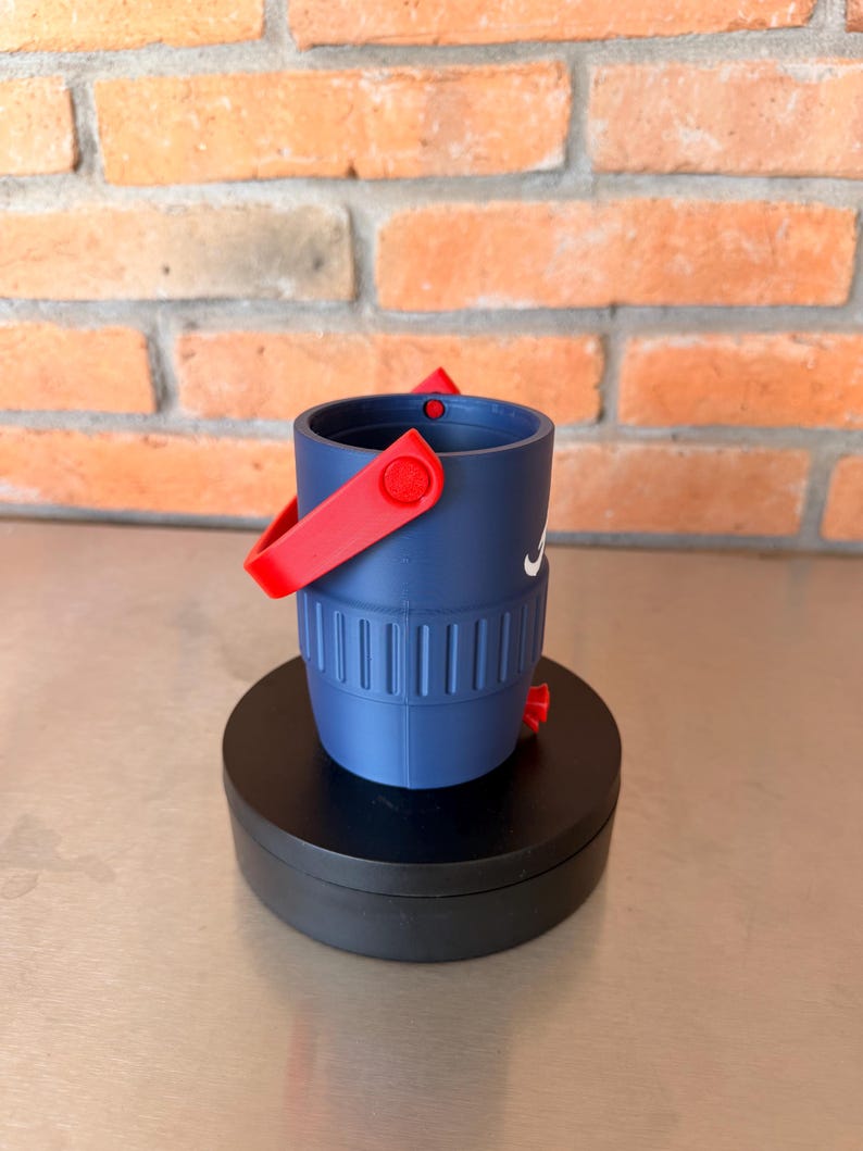 May include: A navy blue bucket-shaped container with a red handle and accents. The container has a white logo on the side and is sitting on a black circular base. The background is a brick wall.