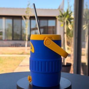 May include: A blue and yellow insulated tumbler with a stainless steel straw. The tumbler has a yellow handle and lid, with a blue body. The tumbler is designed to keep drinks cold. The tumbler is sitting on a black surface.