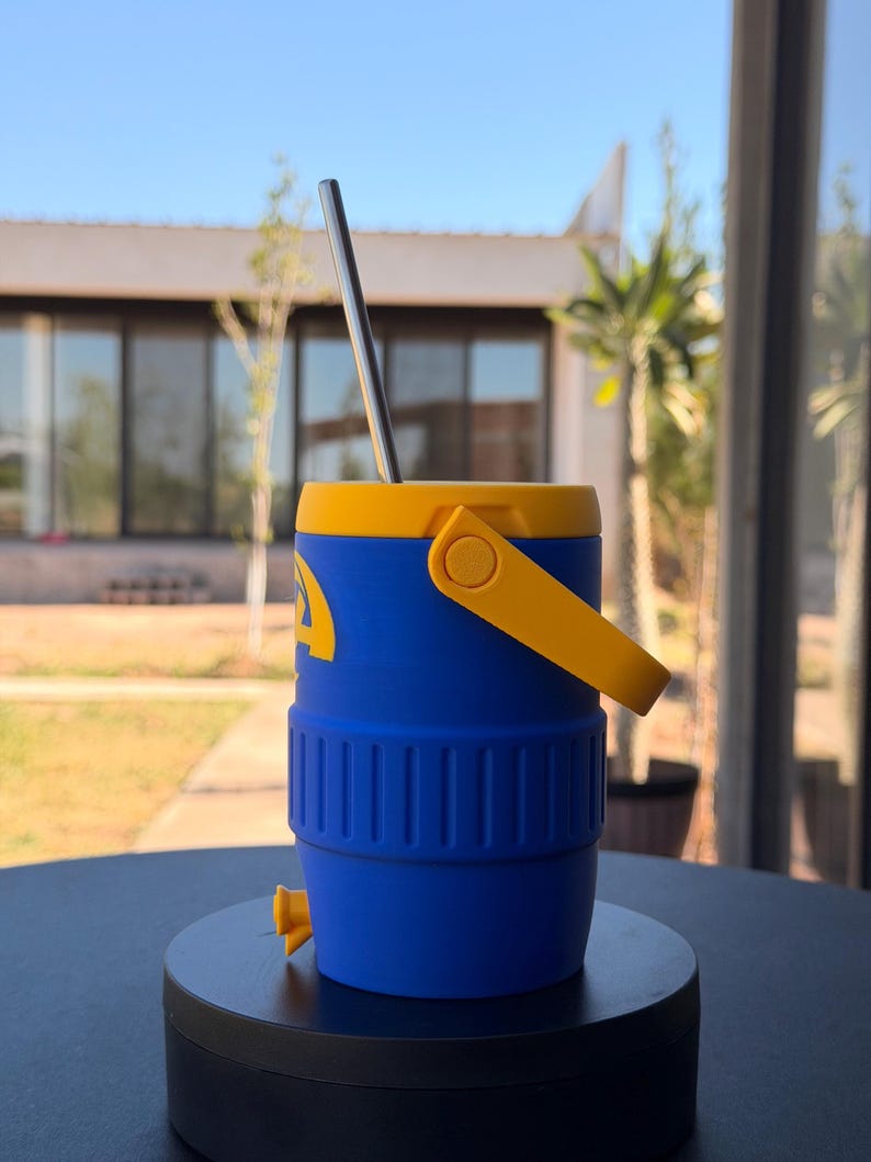 May include: A blue and yellow insulated tumbler with a stainless steel straw. The tumbler has a yellow handle and lid, with a blue body. The tumbler is designed to keep drinks cold. The tumbler is sitting on a black surface.