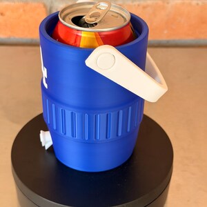 May include: A blue can cooler with a white handle holds a red and yellow can. The cooler is cylindrical with a white logo and sits on a black rotating stand. The background is a brick wall.