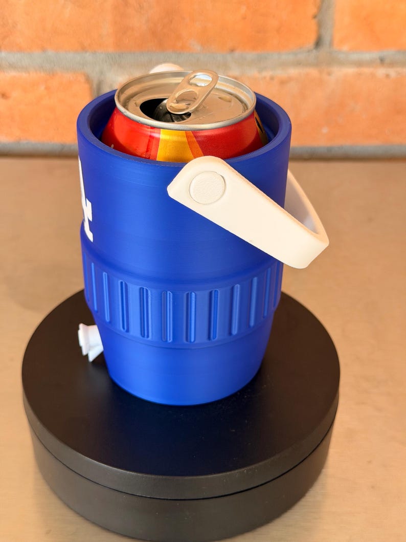 May include: A blue can cooler with a white handle holds a red and yellow can. The cooler is cylindrical with a white logo and sits on a black rotating stand. The background is a brick wall.