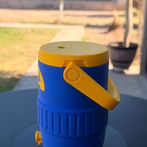 May include: A blue and yellow water cooler with a handle. The cooler has a yellow lid and handle, with a blue body. It features a spigot and a small hole on the top. The cooler is sitting on a black surface.