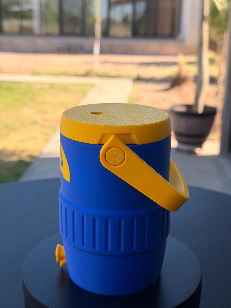 May include: A blue and yellow water cooler with a handle. The cooler has a yellow lid and handle, with a blue body. It features a spigot and a small hole on the top. The cooler is sitting on a black surface.