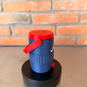 May include: A navy blue and red insulated water jug with a white logo. The jug has a red handle and lid. The jug is cylindrical with vertical grooves and sits on a black base.