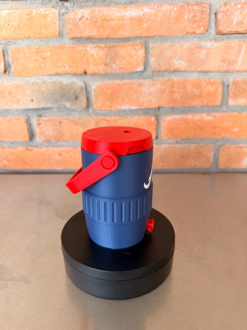 May include: A navy blue and red insulated water jug with a white logo. The jug has a red handle and lid. The jug is cylindrical with vertical grooves and sits on a black base.