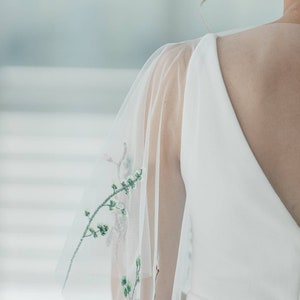 May include: A white wedding dress with a sheer overlay featuring delicate green floral embroidery. The dress has a V-neckline and a fitted bodice.