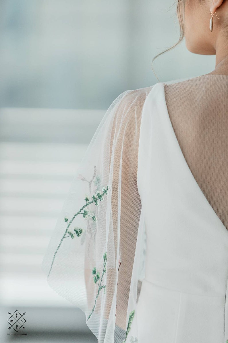 May include: A white wedding dress with a sheer overlay featuring delicate green floral embroidery. The dress has a V-neckline and a fitted bodice.