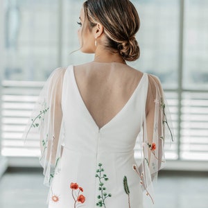 May include: A white wedding dress with a deep V-back and sheer floral embroidered sleeves. The dress has a fitted bodice and a flowing skirt. The embroidery features red, green, and white flowers.