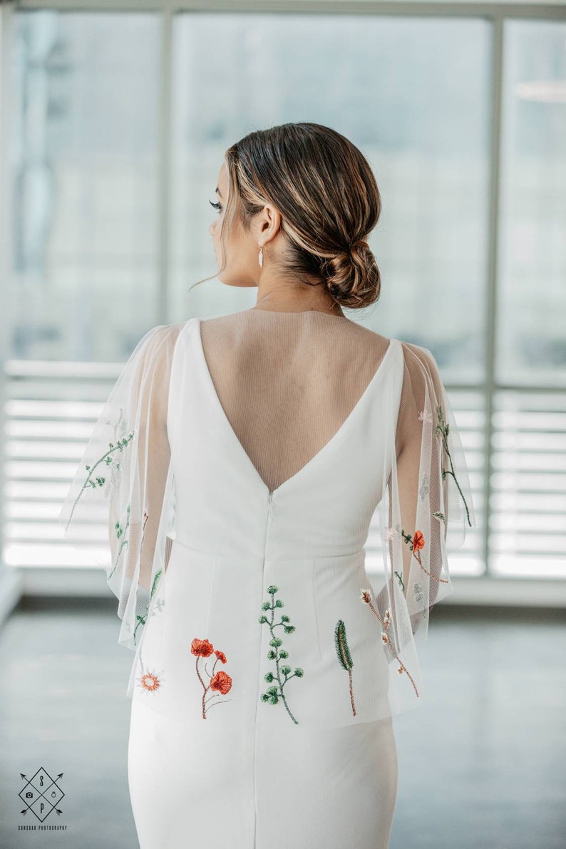 May include: A white wedding dress with a deep V-back and sheer floral embroidered sleeves. The dress has a fitted bodice and a flowing skirt. The embroidery features red, green, and white flowers.