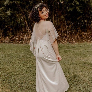 May include: A woman wearing a white dress with a sheer overlay embroidered with flowers and greenery. She is wearing glasses and has curly hair.