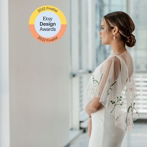 May include: A woman in a white dress with a sheer floral embroidered overlay stands in front of a window. The window is behind her and out of focus. A sticker on the wall reads "2022 Finalist Etsy Design Awards 2022 Finalist".