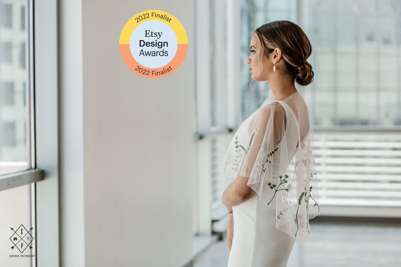May include: A woman in a white dress with a sheer floral embroidered overlay stands in front of a window. The window is behind her and out of focus. A sticker on the wall reads "2022 Finalist Etsy Design Awards 2022 Finalist".