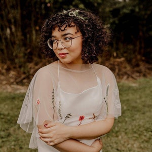 May include: A woman wearing a white dress with floral embroidery and a sheer overlay. She has curly brown hair and is wearing round glasses. She is standing in a grassy field.