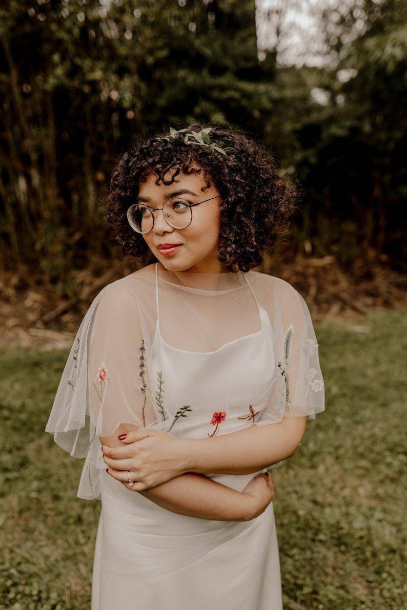 May include: A woman wearing a white dress with floral embroidery and a sheer overlay. She has curly brown hair and is wearing round glasses. She is standing in a grassy field.