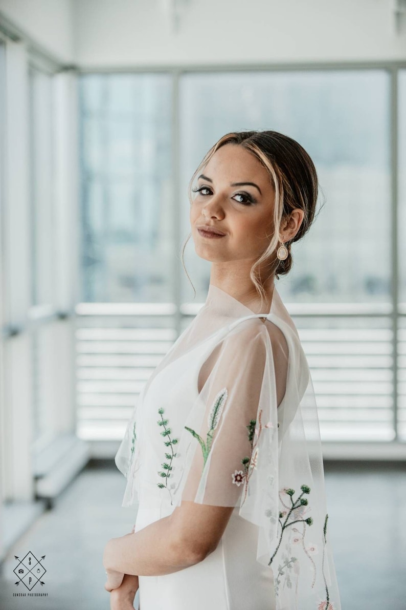 May include: A woman in a white dress with floral embroidery on the sleeves. She is standing in front of a window with a white background.