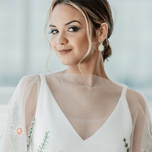 May include: A woman wearing a white dress with floral embroidery. The dress has a sheer overlay and a V-neckline. She is wearing delicate earrings with a green gemstone.