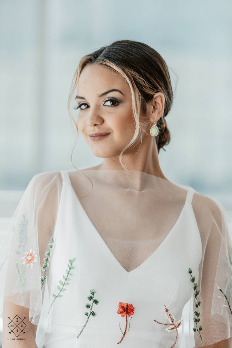 May include: A woman wearing a white dress with floral embroidery. The dress has a sheer overlay and a V-neckline. She is wearing delicate earrings with a green gemstone.