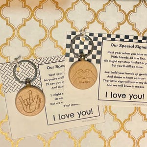 May include: Two wooden keychains with silver keyrings and engraved designs. One keychain features a hand making the "I love you" sign, the other shows hands forming a heart. Each keychain has the text "2024-2025".