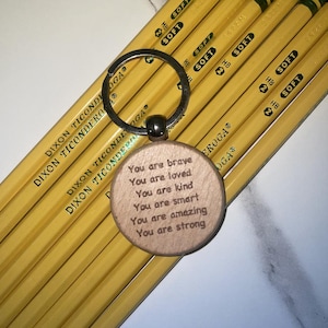 Puede incluir: Un llavero de madera con un anillo plateado y un colgante redondo. El colgante tiene el texto grabado "You are brave, You are loved, You are kind, You are smart, You are amazing, You are strong." Lápices amarillos en el fondo.