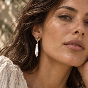 May include: A drop earring featuring a textured, irregular gold-tone metal stud at the top, connected to a long, slender, white baroque pearl pendant with an organic, elongated shape and a lustrous, uneven surface finish.