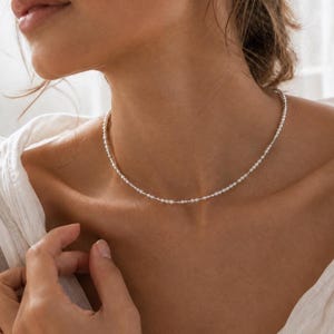 Puede incluir: Un delicado collar tipo gargantilla que presenta una sola hilera de diminutas cuentas blancas uniformes, espaciadas equitativamente a lo largo de una fina cadena metálica. El collar se ajusta a la base del cuello con un diseño sutil y refinado.