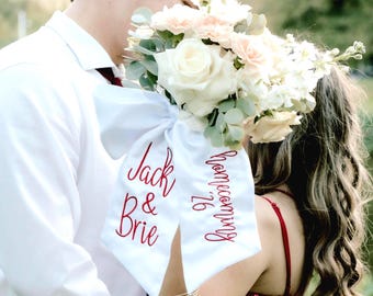 May include: A white satin ribbon tied around a bouquet of white and peach roses and greenery. The ribbon features red embroidered text that reads Jack & Brie on one side and Homecoming '26 on the other side of the fabric tails.