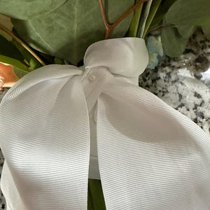 Puede incluir: Una cinta de grosgrain blanca atada en un lazo grande, con un peque&ntilde;o detalle de perla en el centro. La cinta envuelve los tallos de un ramo que contiene hojas de eucalipto verdes y flores claras, con un bordado azul visible.