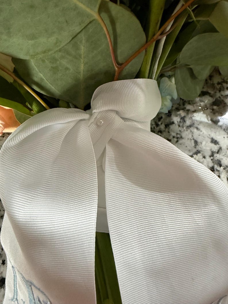 Puede incluir: Una cinta de grosgrain blanca atada en un lazo grande, con un peque&ntilde;o detalle de perla en el centro. La cinta envuelve los tallos de un ramo que contiene hojas de eucalipto verdes y flores claras, con un bordado azul visible.