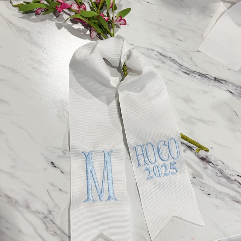 Puede incluir: Una banda de tela blanca con bordado en azul claro. El lado izquierdo muestra la letra M, mientras que el lado derecho presenta el texto HOCO 2025. La banda tiene una textura acanalada y termina en una forma puntiaguda de cola de golondrina.