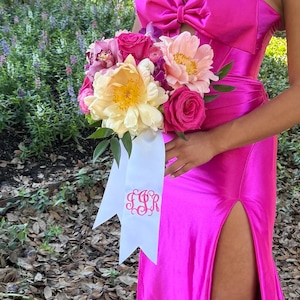 Puede incluir: Un vestido de sat&eacute;n rosa brillante con abertura lateral y un lazo grande en el corpi&ntilde;o, junto a un ramo de flores color crema, rosa y morado. Del ramo cuelga una cinta blanca con un monograma bordado en rosa con las letras JSR.