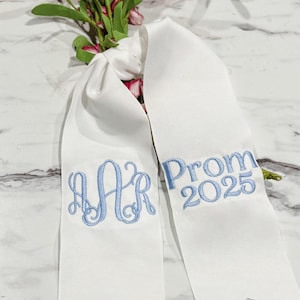 Puede incluir: Cinta de satén blanca con bordado azul. La cinta dice "Prom 2025" y tiene un monograma "AR" en el otro lado.