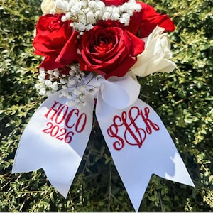 May include: A floral bouquet featuring deep red and white roses accented with small white baby's breath flowers. A white ribbon is tied at the base, embroidered with red text that reads HOCO 2026 on one side and a stylized monogram SJK on the other.