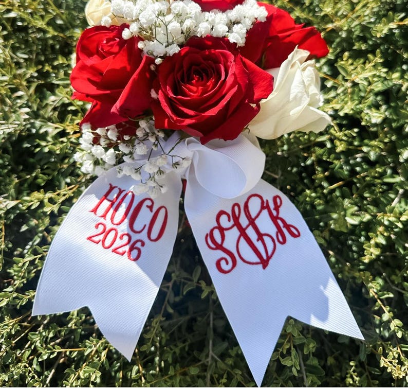 Puede incluir: Un ramo de flores con rosas de color rojo intenso y blanco, acentuado con peque&ntilde;as flores blancas de paniculata. Una cinta blanca est&aacute; atada en la base, bordada con texto rojo que dice HOCO 2026 en un lado y un monograma estilizado SJK en el otro.