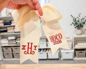 May include: A cream-colored hair bow made of grosgrain ribbon with two long tails. The left tail features a red embroidered monogram with the letters ZHY, and the right tail features red embroidered text reading HOCO 2026.