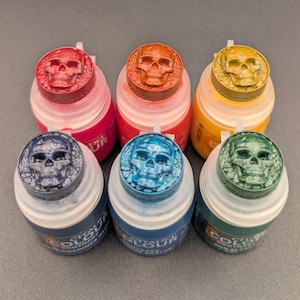 Könnte beinhalten: Sechs Kunststoff-Farbtöpfe in zwei Reihen, jeder mit einem schädelförmigen Deckel in einer anderen Metallic-Farbe: Rot, Orange, Gold, Dunkelblau, Hellblau und Grün. Die durchscheinenden Flaschen enthalten Citadel Colour-Farbe.