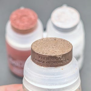 May include: Three small, translucent plastic bottles with round, flat caps in metallic copper, silver, and bronze. The bronze cap features the embossed text "TECHNICAL FORGE OF MEDUSA" on its side. Each bottle has a matching colored base.