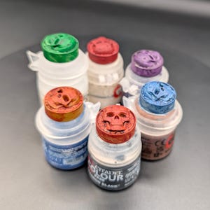 May include: A collection of six Citadel Colour paint pots with translucent plastic bodies and colored lids. Each lid features a molded skull design. The pots contain various paint colors including green, red, purple, blue, and black, with visible text.