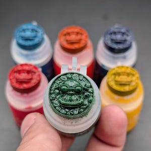 Puede incluir: Una colección de seis pequeños botes de pintura de plástico con cuerpos translúcidos y tapas de colores. Cada tapa presenta un relieve circular de una cara de criatura estilizada rodeada por una textura de piedra. Los colores incluyen azul, naranja, azul oscuro, rojo, amarillo y una tapa verde prominente en primer plano.