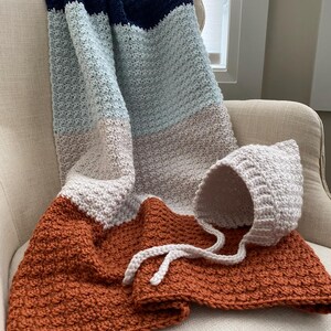 May include: A handmade crochet blanket and bonnet set. The blanket features stripes of navy blue, light blue, gray, and rust orange. The bonnet is light gray with white ties. The set is displayed on a cream-colored chair.